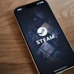 Telefon z aplikacją Steam, gdzie widać logo i okładki gier. W tle delikatna **steam cloud** unosząca się nad ekranem.