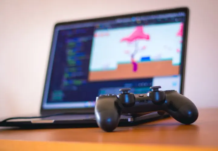 Gamepad do PS4 leży przed laptopem z kodem i grą na ekranie. Dowiedz się, jak podłączyć PS4 do laptopa, by grać gdziekolwiek.