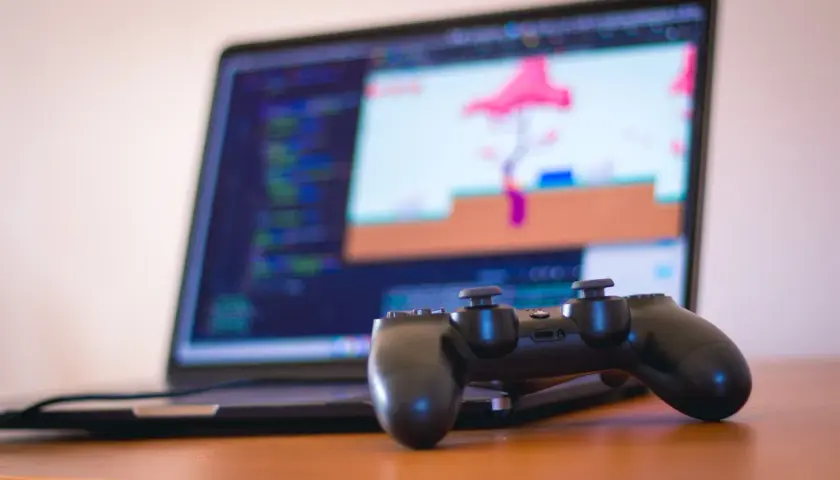 Gamepad do PS4 leży przed laptopem z kodem i grą na ekranie. Dowiedz się, jak podłączyć PS4 do laptopa, by grać gdziekolwiek.