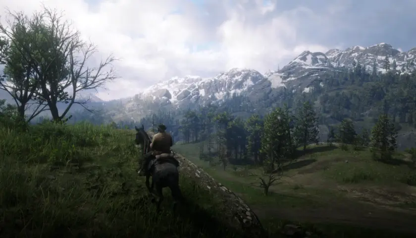 Jazda konno przez górzyste tereny w Red Dead Redemption 2 PC. Śnieżne szczyty gór i zielone lasy tworzą zapierający dech w piersiach krajobraz.