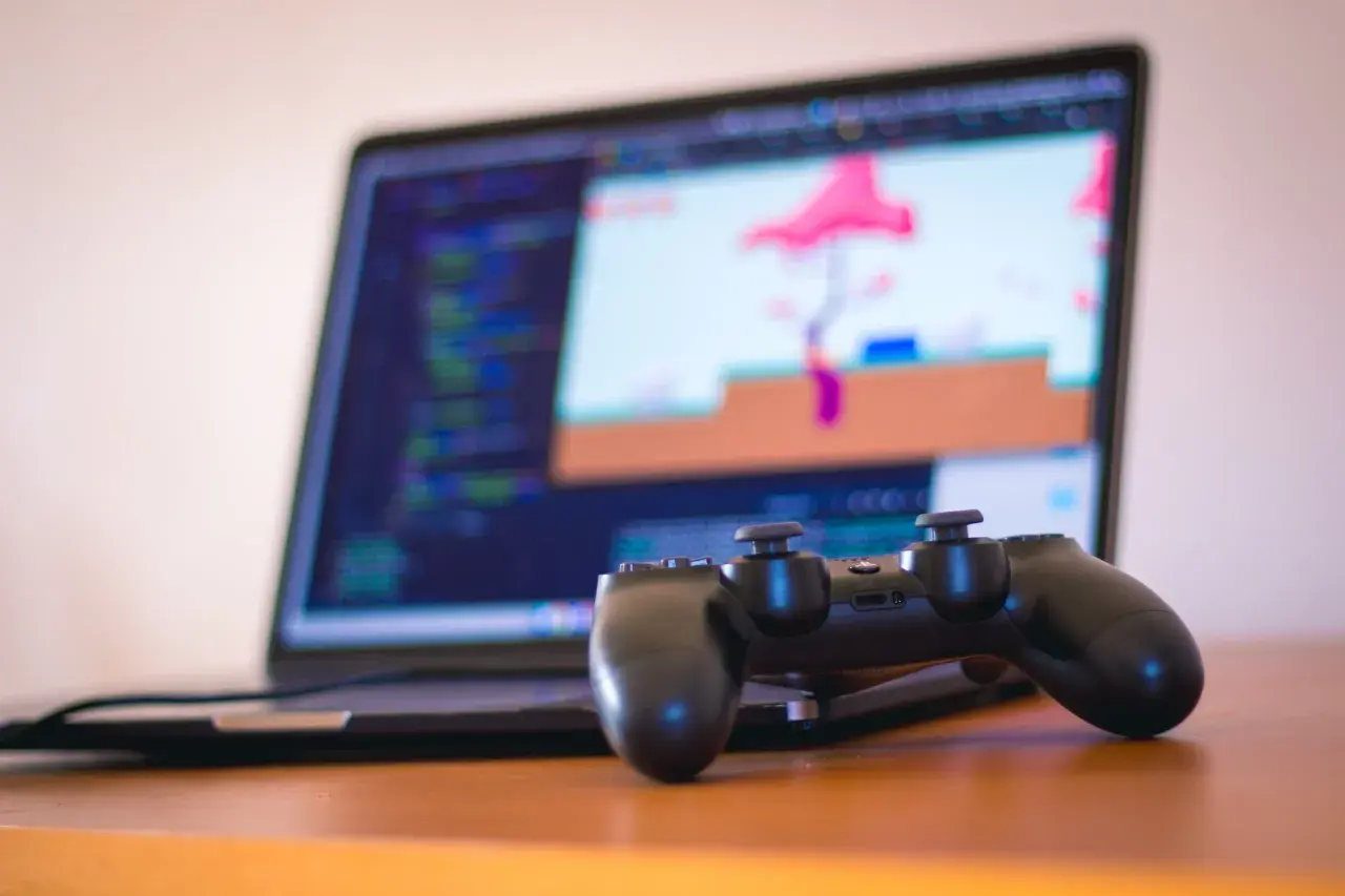 Gamepad do PS4 leży przed laptopem z kodem i grą na ekranie. Dowiedz się, jak podłączyć PS4 do laptopa, by grać gdziekolwiek.