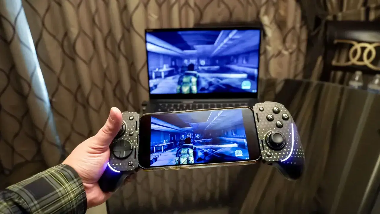Gracz trzyma smartfon z kontrolerem, grając w grę dzięki technologii remote play. Na ekranie laptopa w tle widać ten sam obraz.